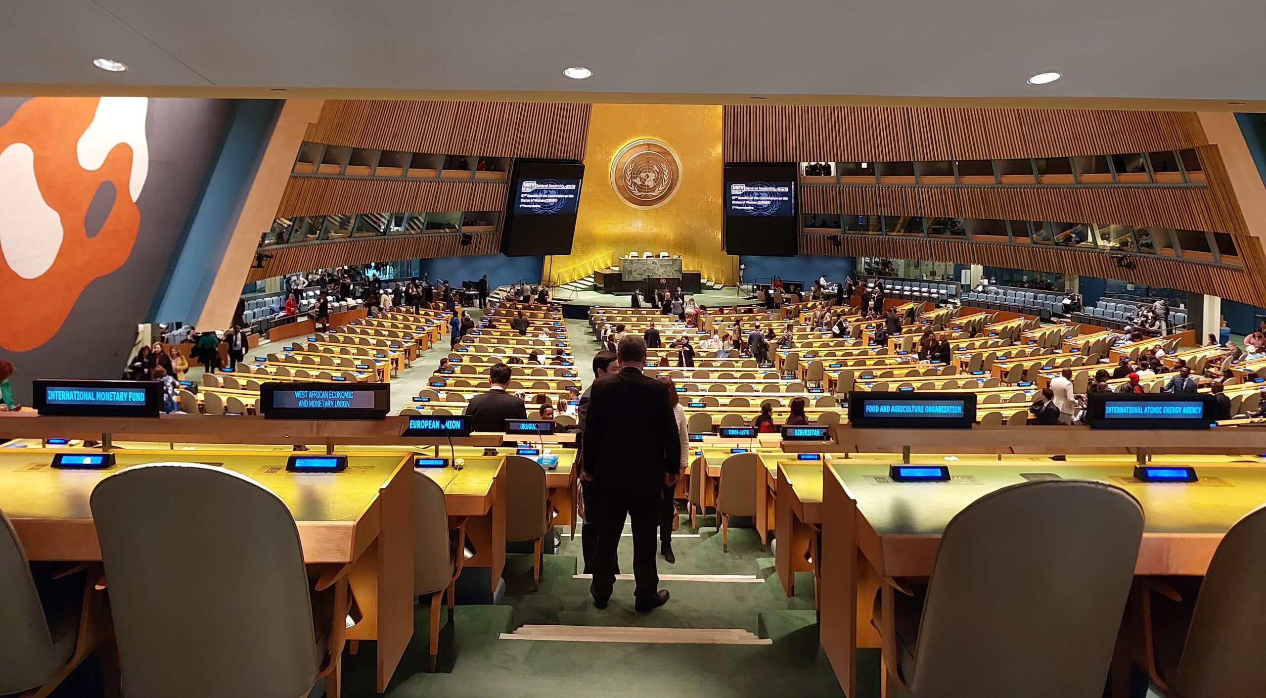 Fotografía del salón principal de asambleas de las Naciones Unidas, el cual es de forma circular. Al fondo, al centro y en lo alto está el logo de la ONU hecho en un metal, de color cobre; está iluminado por una luz blanca que contrasta con el fondo metálico. Además, en lo alto y por los costados hay una serie de ventanas, que es donde están los traductores y traductoras. Bajo el logo, hay un mesón principal levemente más alto, rodeado por pantallas, donde suelen sentarse quienes presiden y moderan la asamblea y luego, hay varias corridas con cientos de sillas y mesones de manera. Cada mesón tiene un letrero digital con el nombre de un país u organización al cual se ha asignado el asiento, además de un micrófono y sistema de sonido. En la sala se ven a algunas personas conversando o de pie durante un receso.