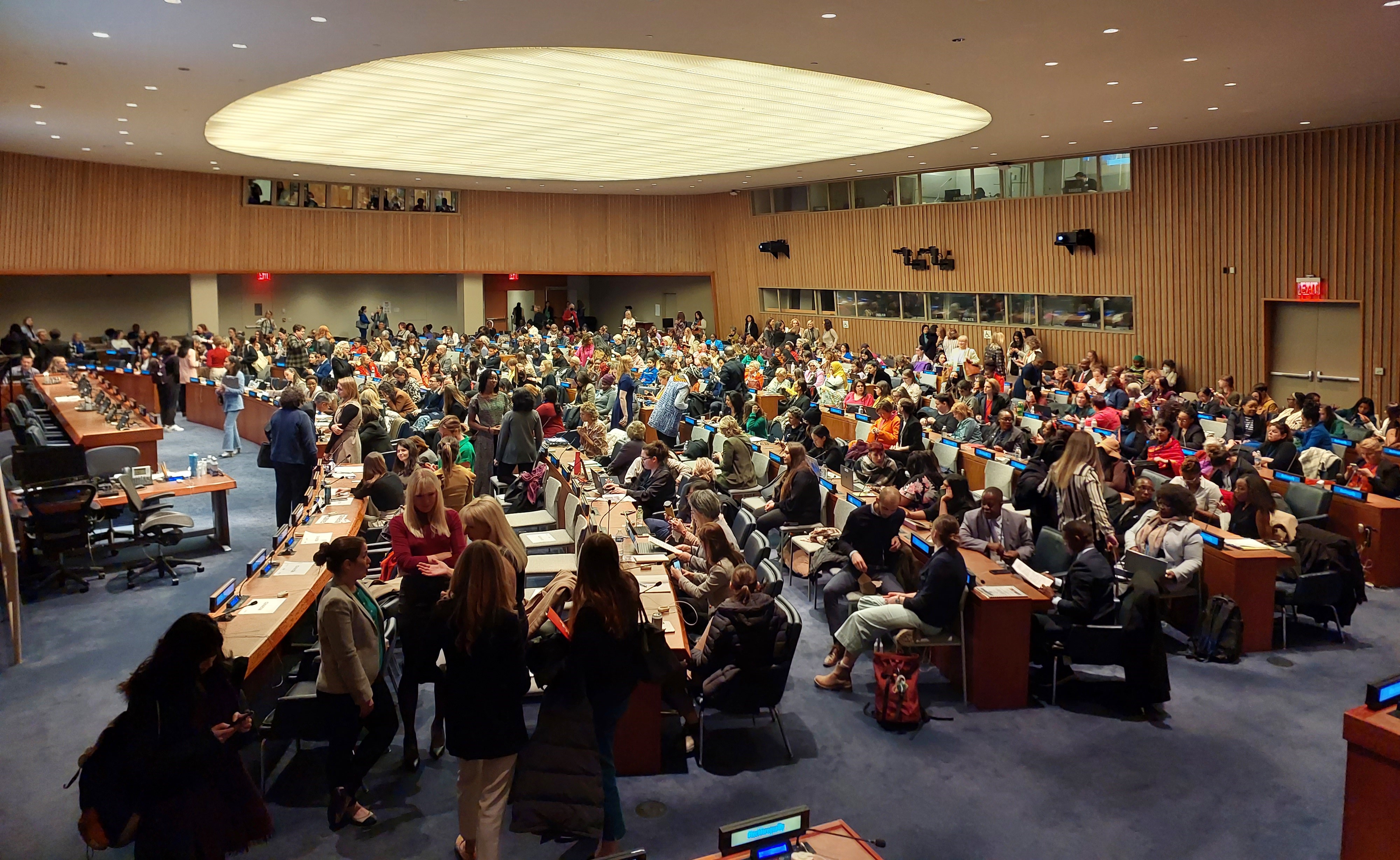 Imagen de una sesión en una sala de conferencias al interior de las Naciones Unidas. Las cientas de sillas y mesas de madera están dispuestas de forma semicircular; son de madera y cada una cuenta con un letrero electrónico que anuncia quién se sienta en cada lugar y un micrófono. Hay cientos de mujeres de todos los países e idiomas sentadas, conversando o de pie por el lugar. Al fondo y en lo alto se ven unas ventanas, que es donde suelen estar los intérpretes y traductores.